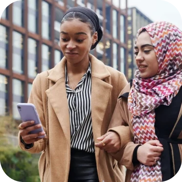 A photograph of two young women walking together outdoors in an urban setting, with one holding and looking at a light purple smartphone.